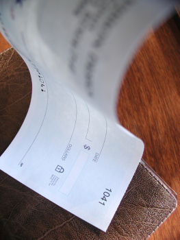 This color photo shows a brown checkbook on a woodgrain desk as a background with a bouncing bank check as the main subject. The bottom of the check is in focus with the top part of the check blurred and slightly in motion to give a "bouncing” appearance. Image is in portrait orientation.